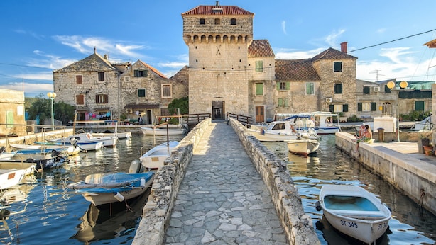 Un bâtiment en pierre doté d’une tour, un sentier en pierre menant à l’eau et plusieurs bateaux amarrés dans une marina.