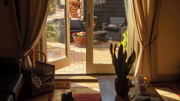 A room with a sliding glass door leading to an outdoor patio area. Inside, there is a wicker basket, a potted plant, a wooden table with a remote control, and a pair of shoes on the floor. - The Old Barn Self Catering Cottage by the Beach, Dog Friendly on Norfolk Coast (Heacham)