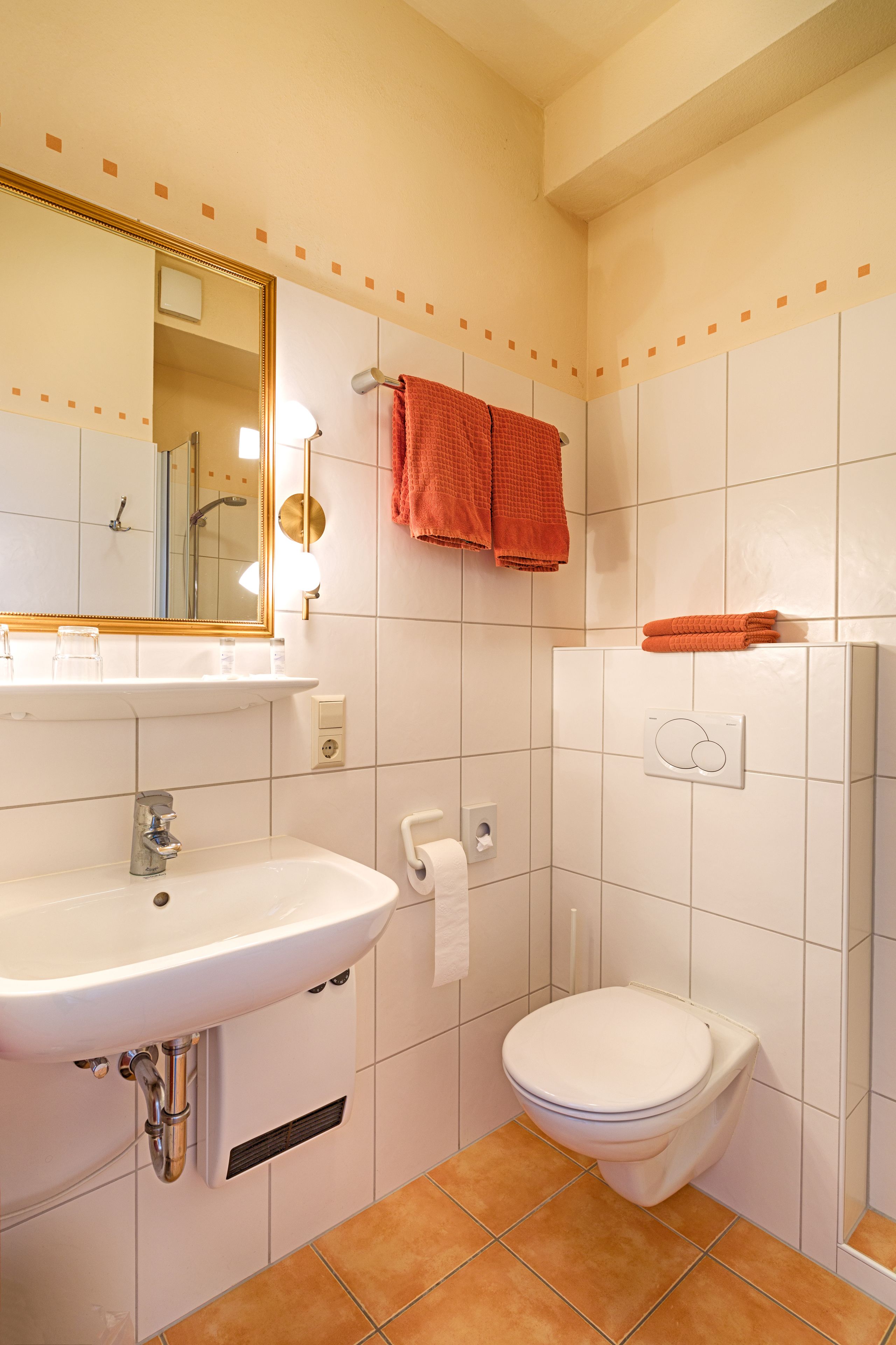 A bathroom with a white sink, mirror, and toilet. There are orange towels hanging on the wall.