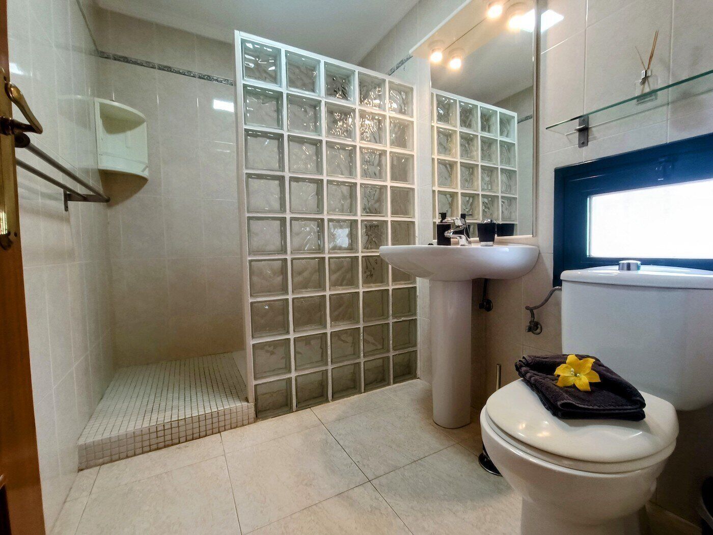 A bathroom with a glass shower enclosure, a pedestal sink, and a toilet with a towel and flower on it.