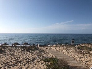Ein Sandstrand mit einem hölzernen Weg zum Meer, einem Rettungsturm und reetgedeckten Sonnenschirmen.