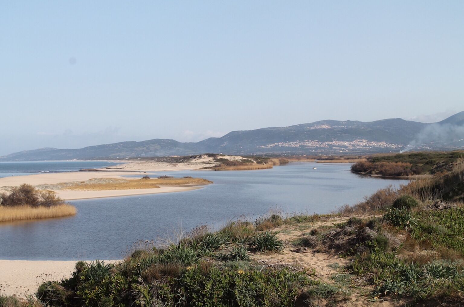 Un paysage côtier avec une plage de sable, une eau calme et des collines à l’horizon.