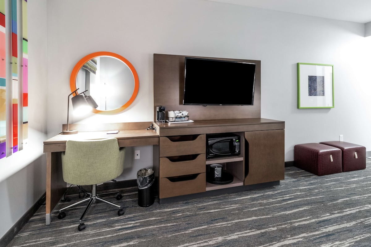A hotel room with a desk, chair, TV, microwave, and storage drawers.