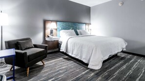 Ein modernes Hotelzimmer mit einem großen Bett, einem dunklen Sessel, einem Schreibtisch und einem Nachttisch mit Lampe.