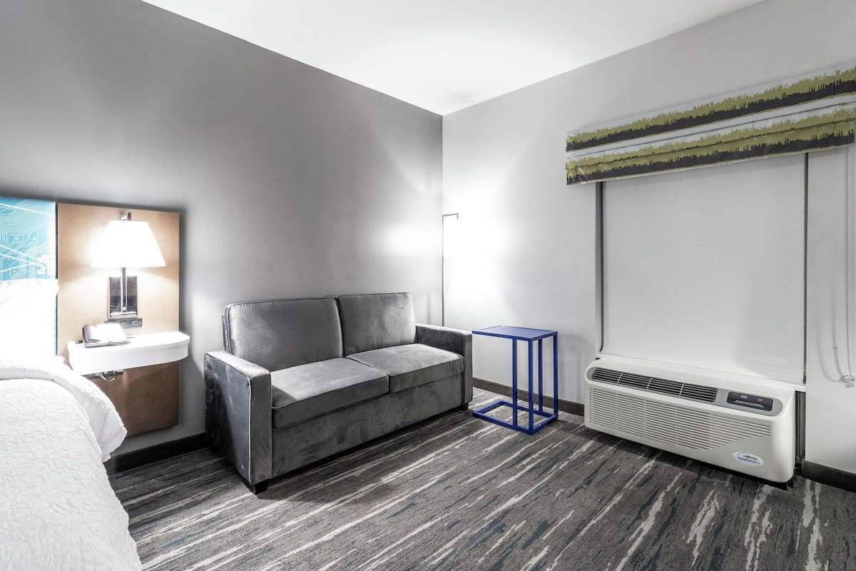A modern hotel room with a grey sofa, a small side table with a lamp, a blue side table, a large air conditioning unit, and a window with blinds.