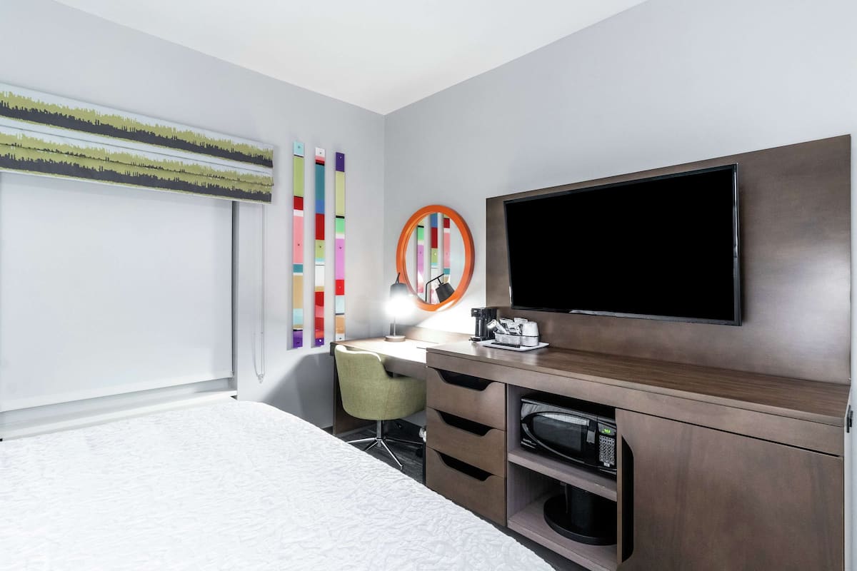 A hotel room with a bed, a desk with a chair, a television, a microwave, and a window with blinds.