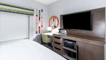 A hotel room with a bed, a desk with a chair, a television, a microwave, and a window with blinds.