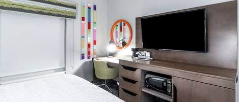 A hotel room with a bed, a desk with a chair, a television, a microwave, and a window with blinds.
