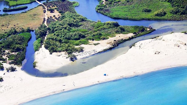 An aerial view of a coastal area with a sandy beach, a river or lagoon, and dense greenery.