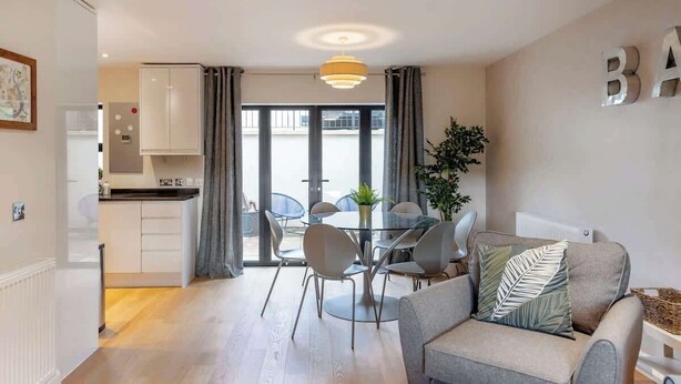 Una sala moderna con un sofá gris, una mesa de comedor de cristal y una cocina al fondo.