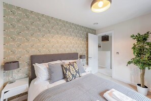 A bedroom with a patterned wallpaper, a bed with multiple pillows, bedside lamps, and a potted plant. - Fantastic 3-bed house in Bath with free parking (BATH)
