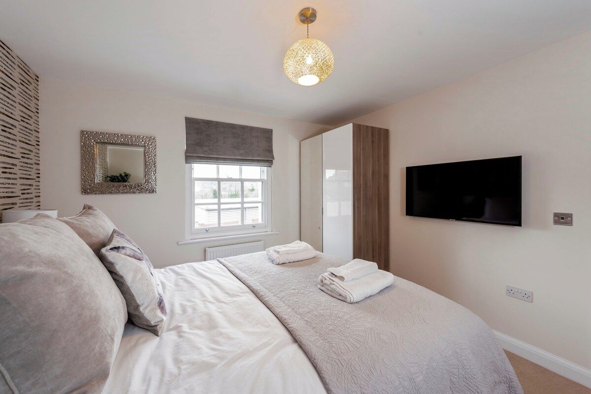 A bedroom with a bed, a television, a window with blinds, a mirror, and a wall-mounted light fixture.