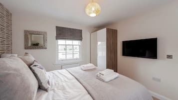 A bedroom with a bed, a television, a window with blinds, a mirror, and a wall-mounted light fixture.