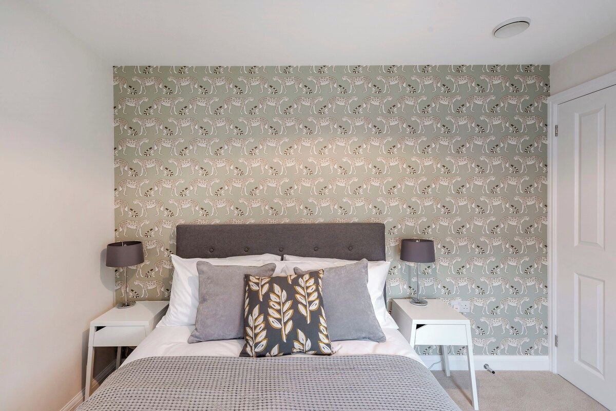 A bedroom with a patterned wallpaper, a bed with pillows, a bedside table, and a nightstand.