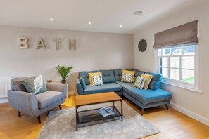 A living room with a grey sofa, a blue sofa, a wooden coffee table, and a grey rug. There is a plant on a side table and a wall-mounted clock. - Fantastic 3-bed house in Bath with free parking (BATH)