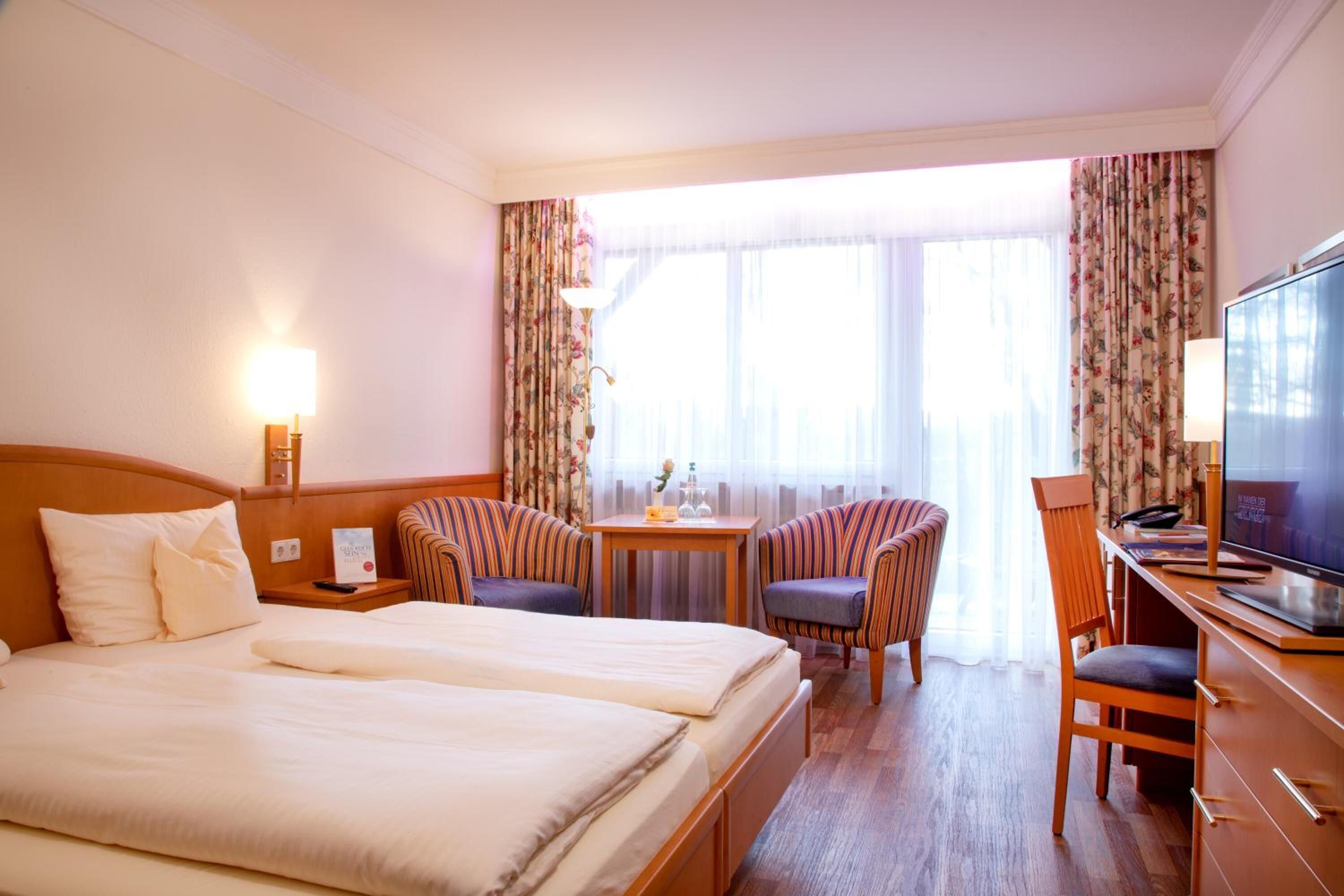 A hotel room with a double bed, two armchairs, a desk with a television, and a small table with a lamp.