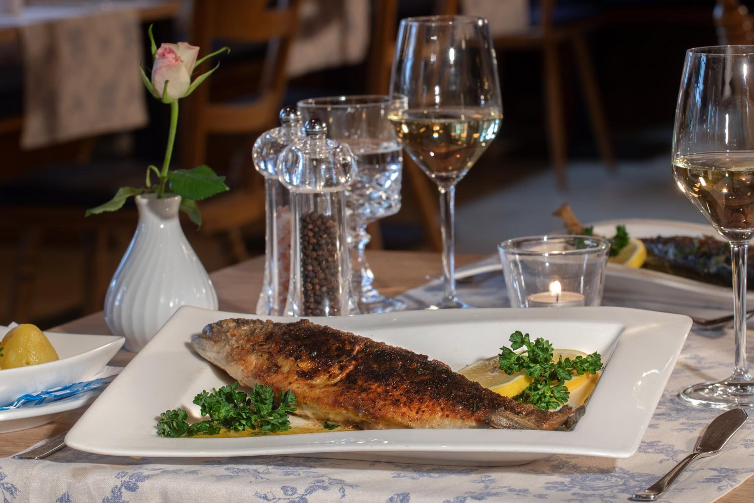 A dining table with a plate of grilled fish, a vase with a rose, salt and pepper shakers, and wine glasses.