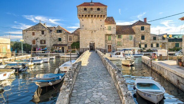 Ein Gebäude aus Stein mit einem Turm, ein Steinweg der zum Wasser führt und mehrere Boote im Hafen vertäut.