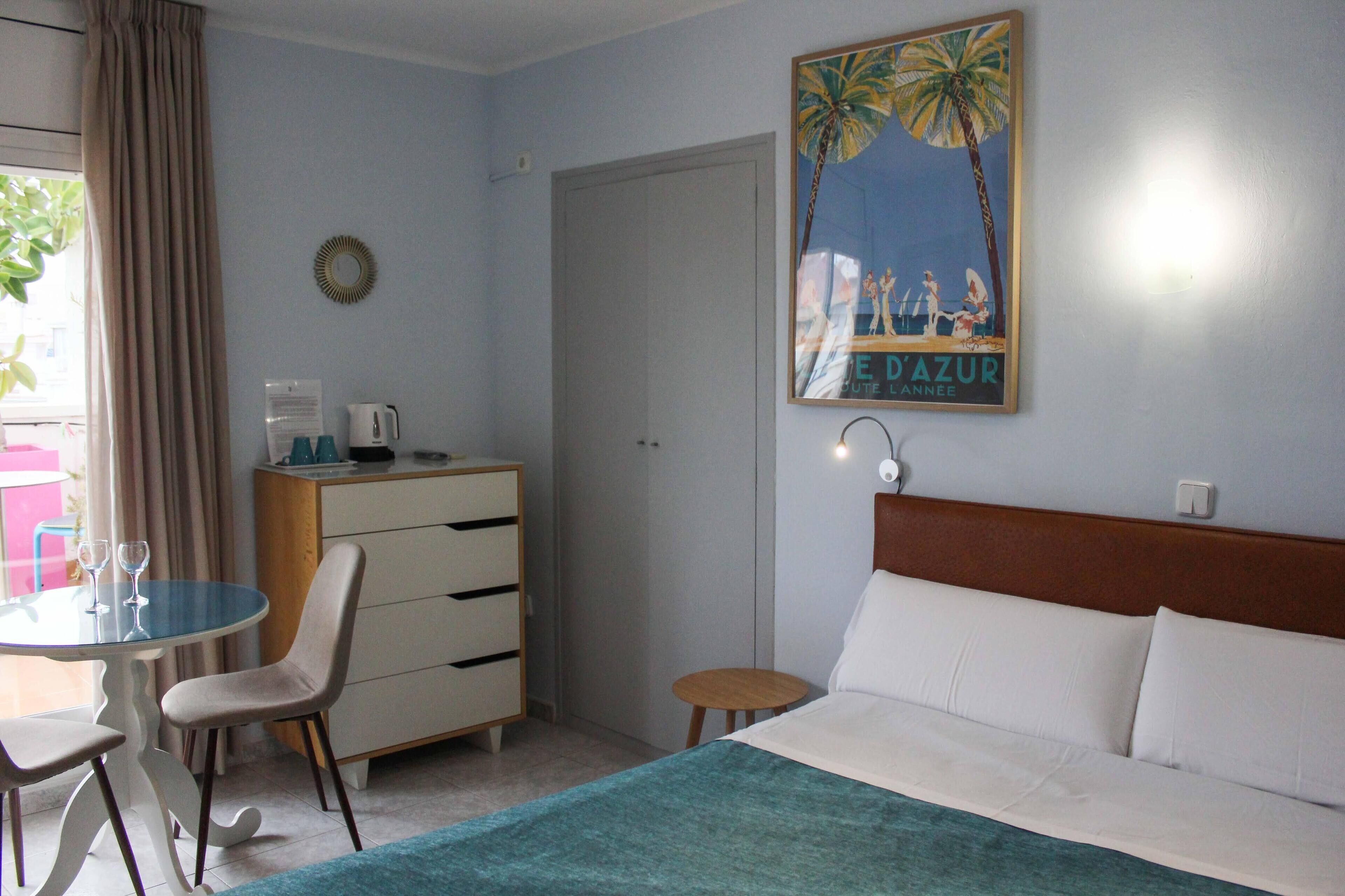 Ein Hotelzimmer mit einem Bett, einem Schreibtisch mit Stuhl, einem kleinen Tisch mit zwei Gläsern, einem Schminktisch und einer Wandlampe.