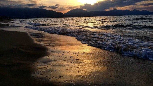 Una playa con olas al atardecer, montañas al fondo y cielo despejado.