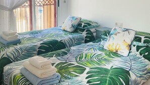 Un dormitorio con una cama cubierta por una colcha con estampado de hojas tropicales, un cuadro de pájaros en la pared y una ventana con cortina.