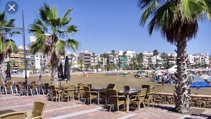 Un área de estar al aire libre con sillas y mesas de mimbre, palmeras y vista a la playa y edificios al fondo.