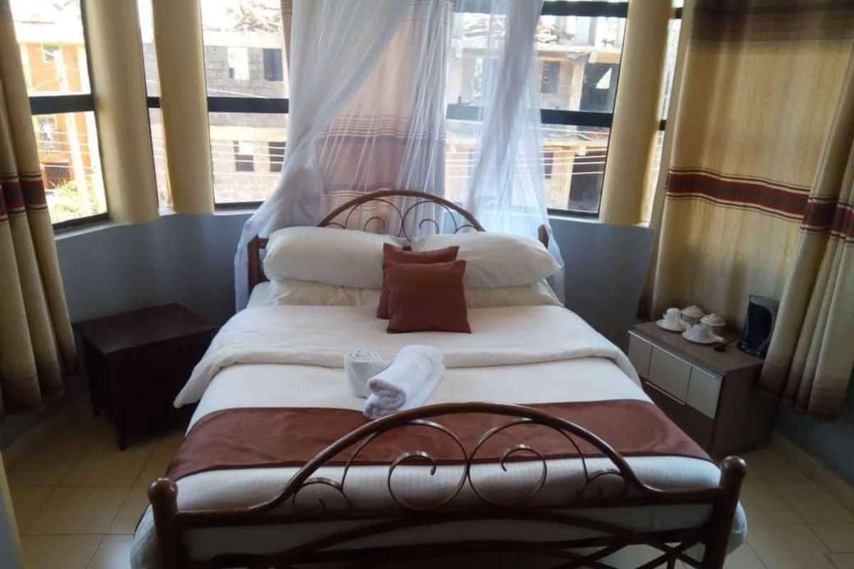 A neatly made bed with a canopy, a bedside table with a cup and saucer, and a view of buildings through large windows.