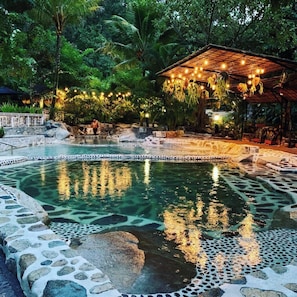 A pool with clear water, surrounded by rocks and plants, and a wooden gazebo with string lights. - Casa Bohemia (Campoalegre)
