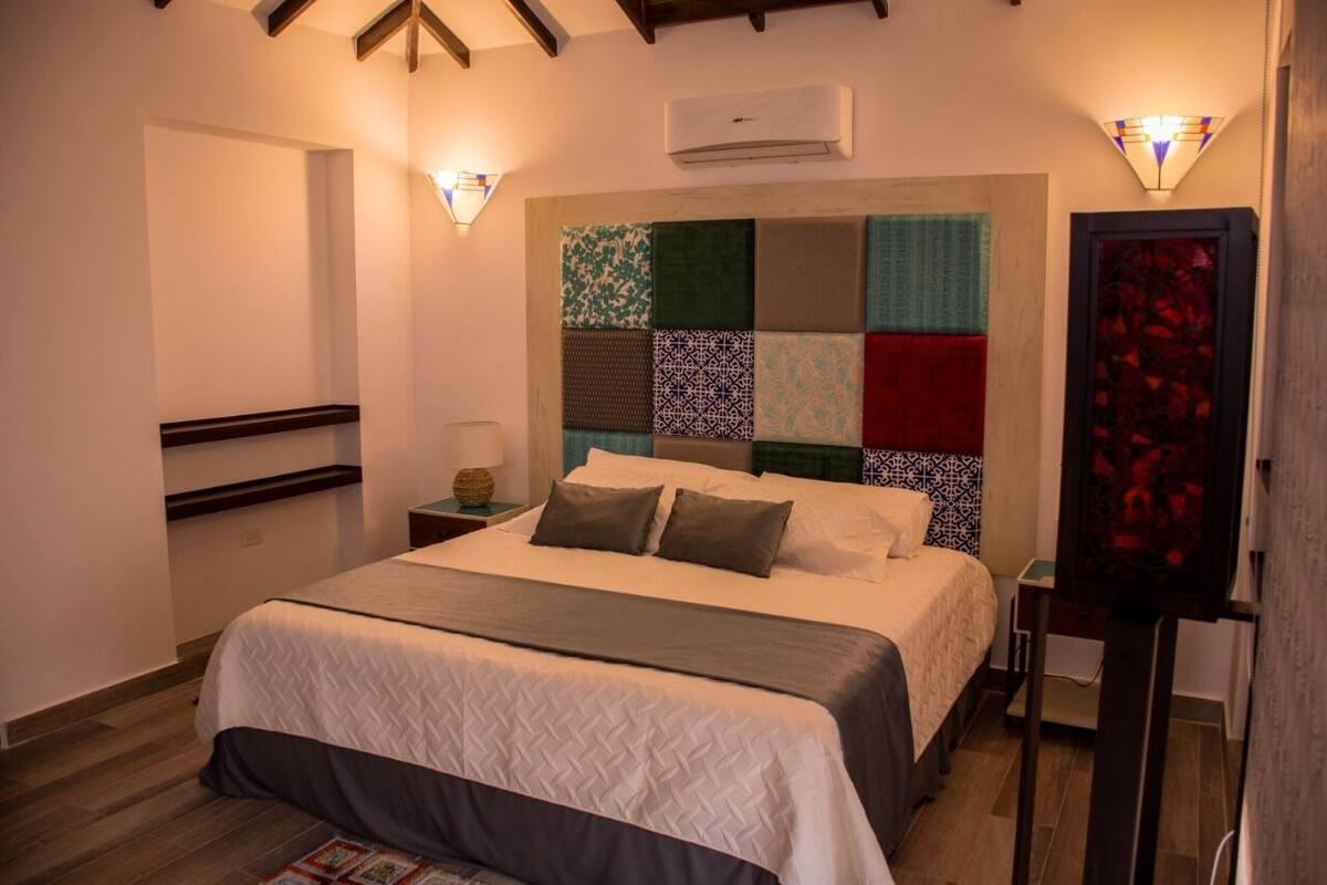 A neatly arranged bedroom with a large bed, a headboard with a patchwork design, a nightstand with a lamp, and a wall-mounted air conditioner.