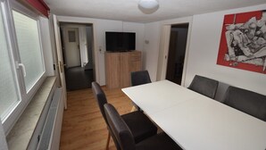Una habitación con una mesa blanca, sillas negras, un televisor sobre un mueble de madera y un ventanal con marco rojo.
