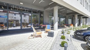 Un edificio moderno con fachada de vidrio, zona de estar exterior con sillones y un coche estacionado cerca.
