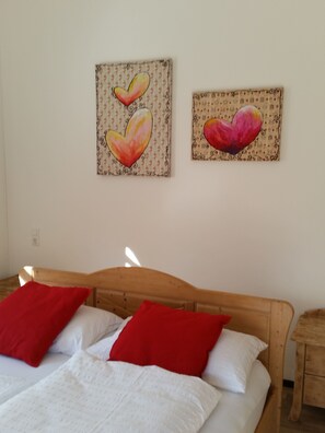 A bedroom with a wooden bed, white bedding, and two red pillows. There are two wall-mounted paintings of hearts. - Masetto Egidio Agritur (Castel Ivano)