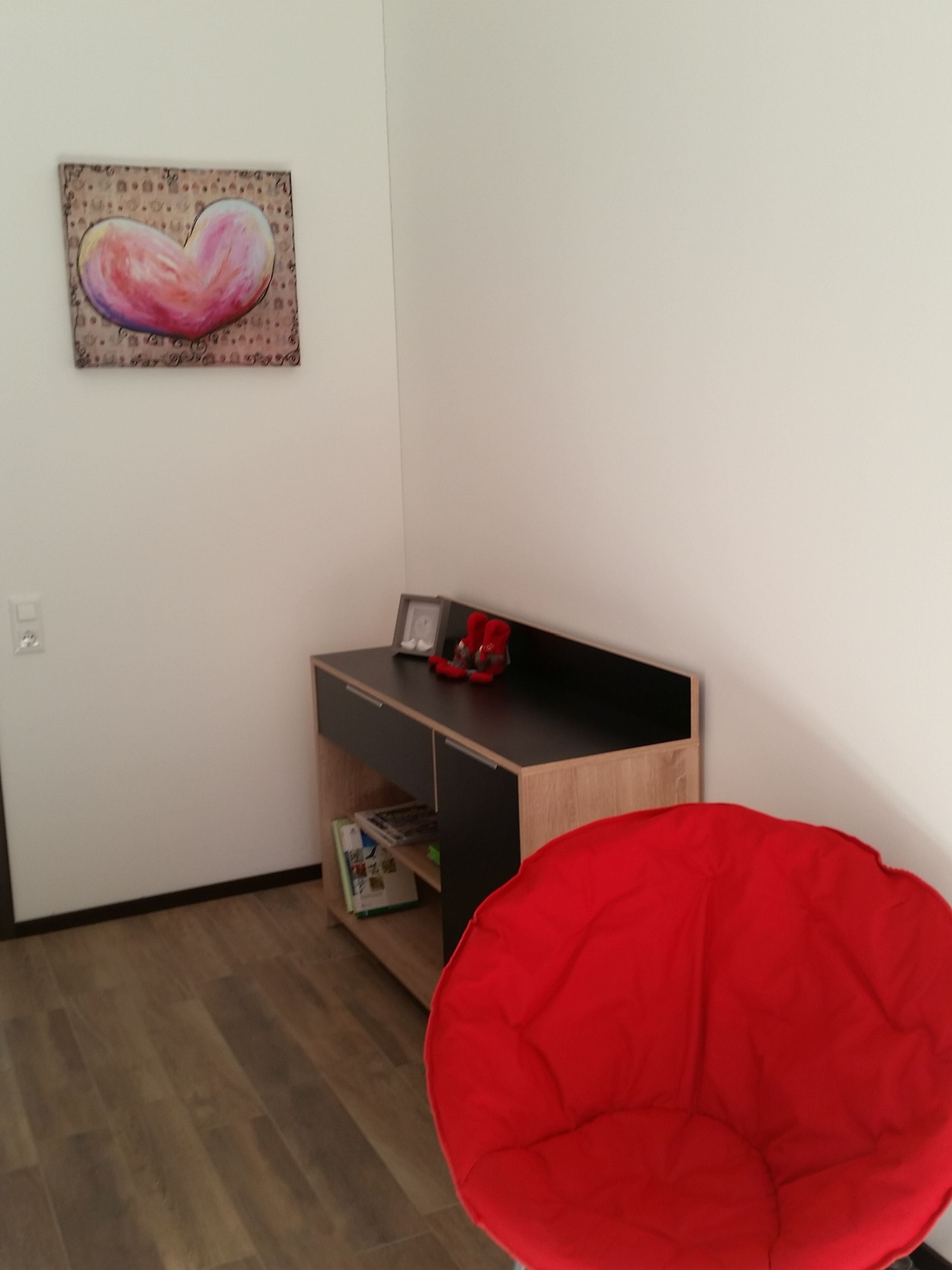 A room with a wooden desk, a red chair, and a heart-shaped painting on the wall.