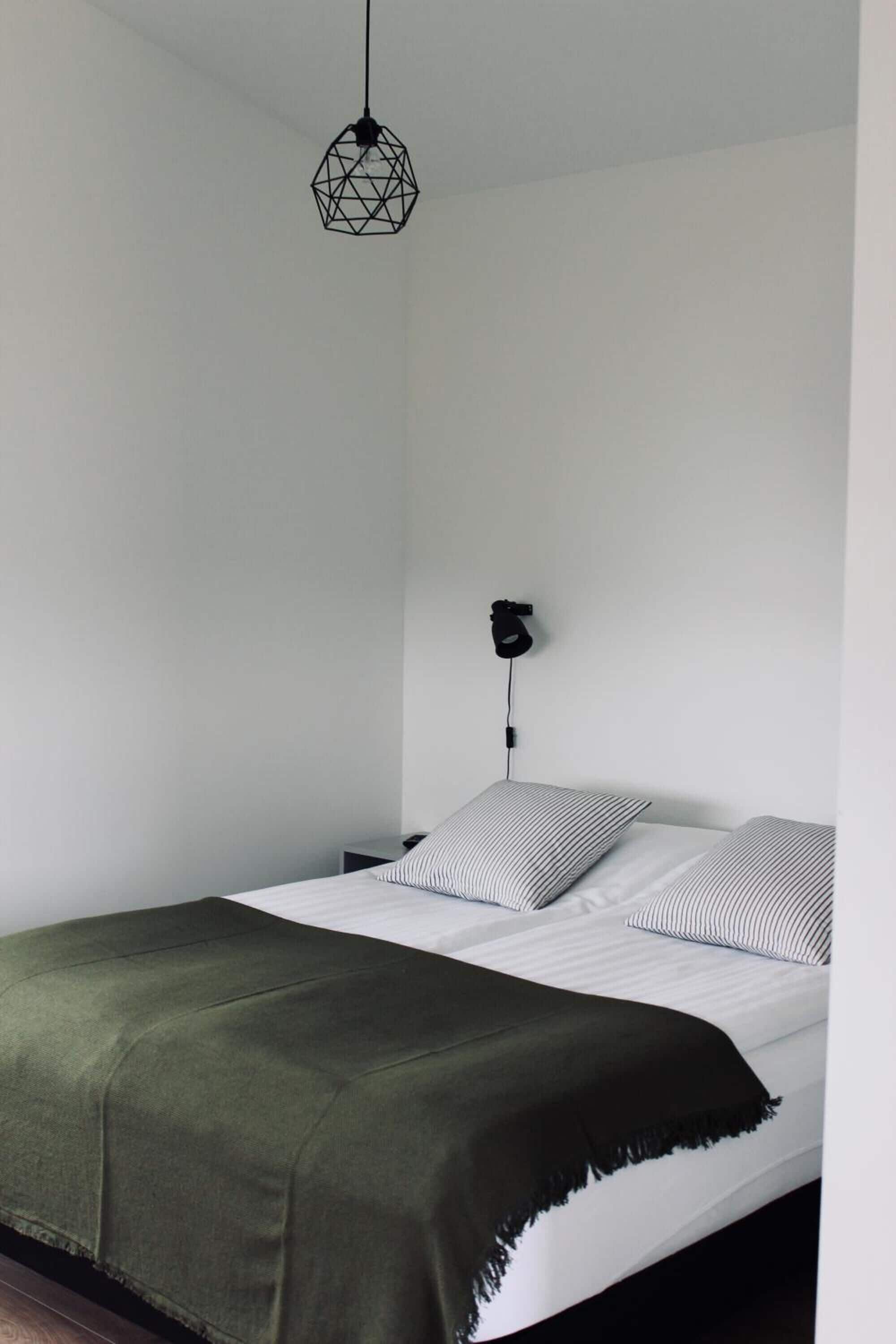 Un lit avec une couverture verte, deux oreillers à rayures et une suspension lumineuse aux motifs géométriques.