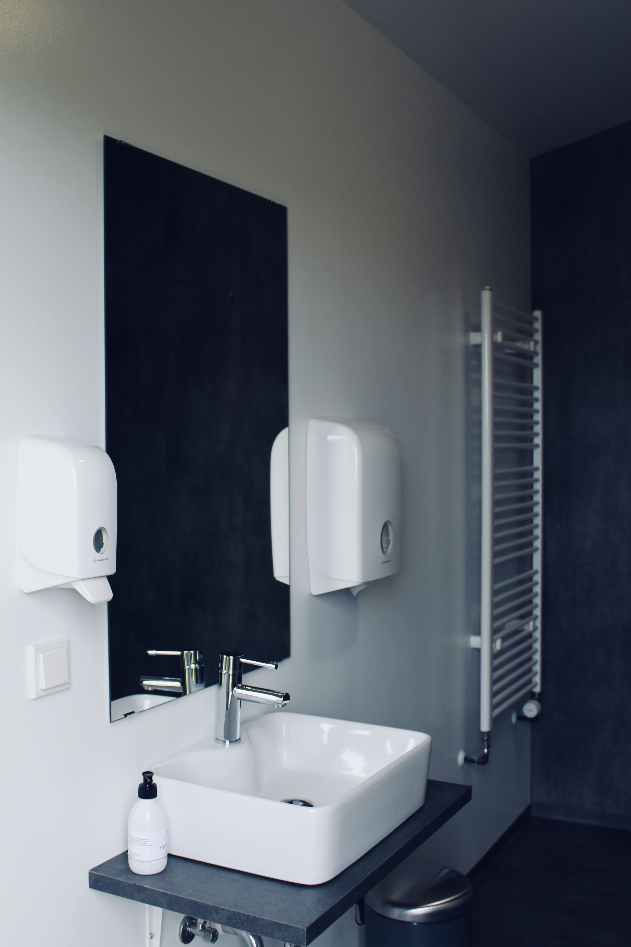 Un baño moderno con lavamanos blanco, encimera negra y un espejo grande. Hay dos dispensadores de jabón en la pared y un calentador de toallas.