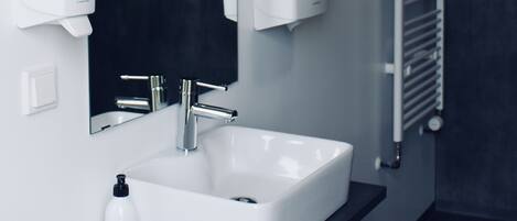 Un baño moderno con lavamanos blanco, encimera negra y un espejo grande. Hay dos dispensadores de jabón en la pared y un calentador de toallas.