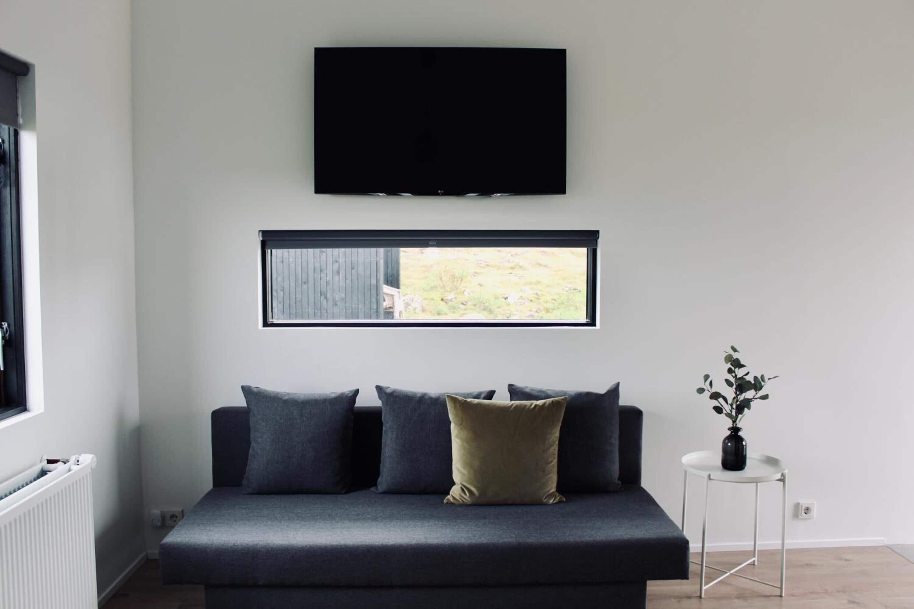 Un salon moderne avec un canapé gris, un téléviseur à écran plat fixé au mur au-dessus, une fenêtre donnant sur un paysage verdoyant et une petite table blanche sur laquelle est posé un vase.