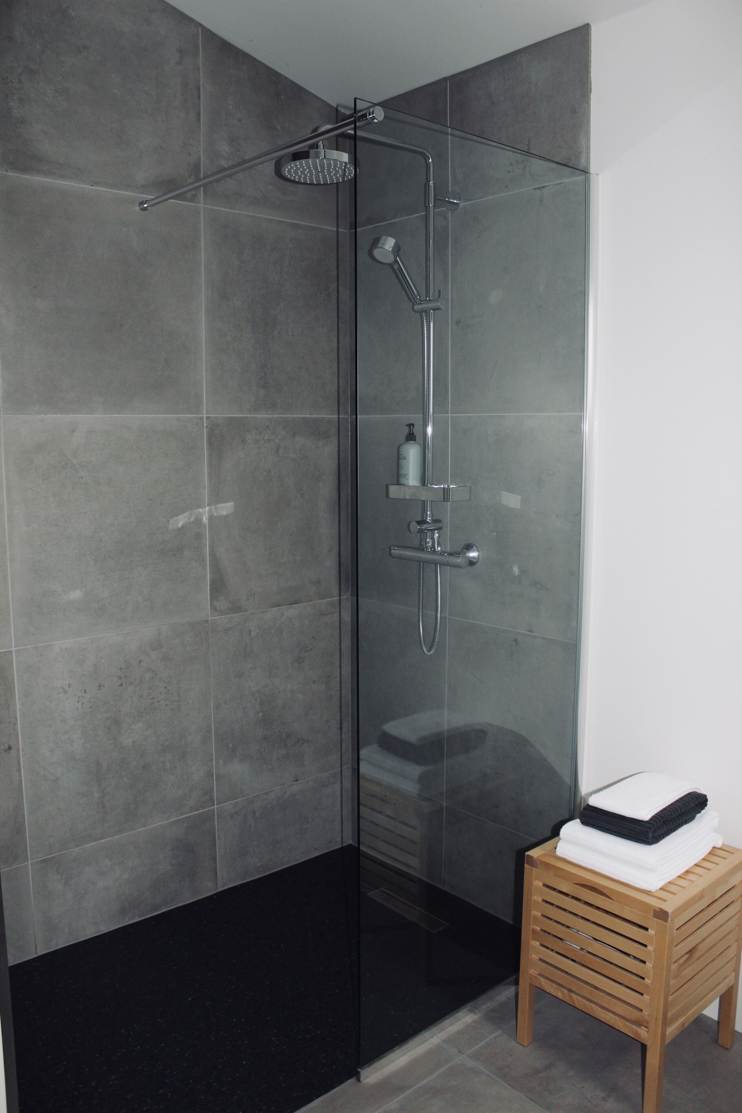 Una doccia moderna con cabina in vetro, uno sgabello di legno con asciugamani e uno scaffale con articoli da toilette.
