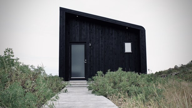 Um edifício moderno preto com uma porta única e uma pequena janela, rodeado por vegetação natural.