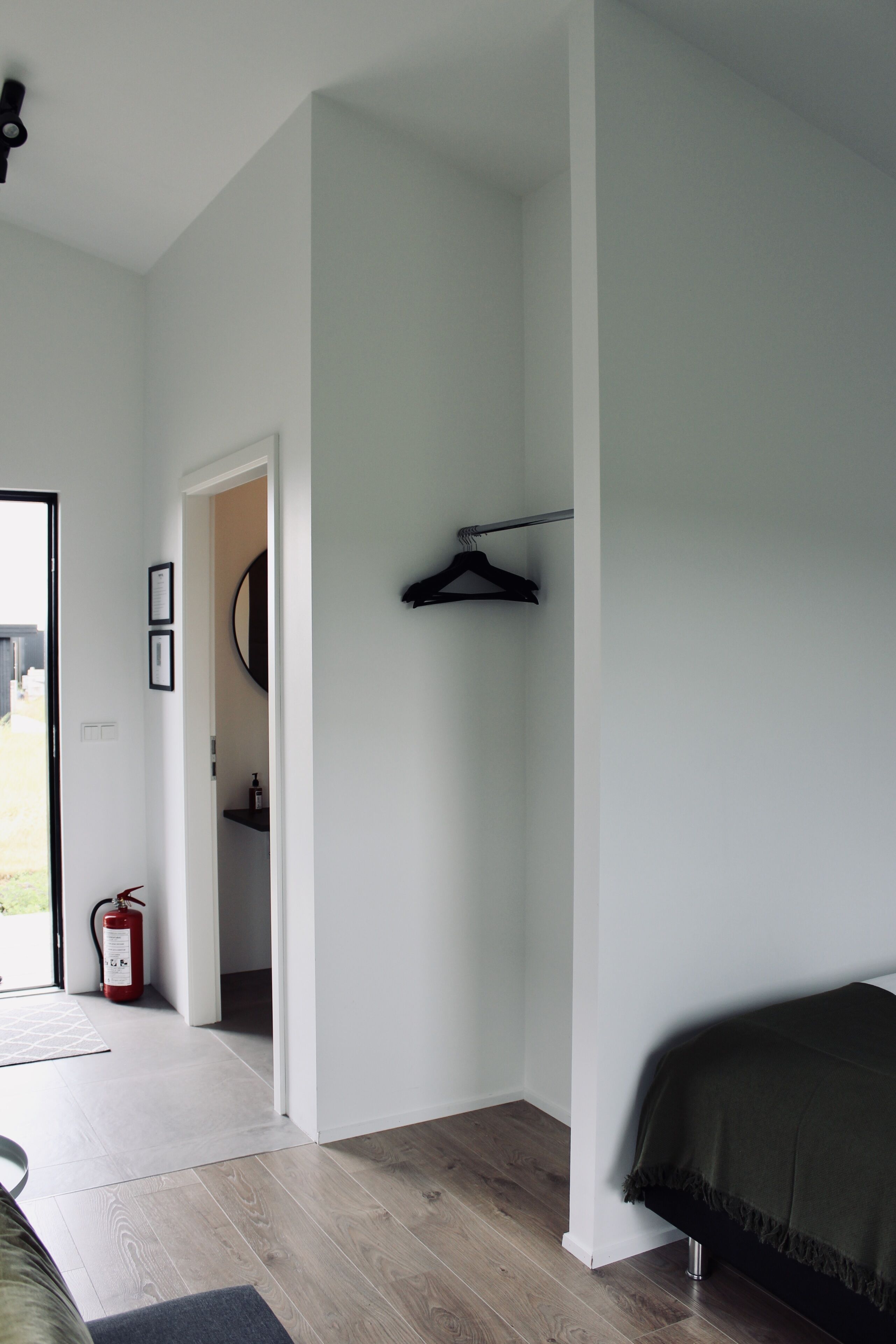 A bedroom with a bed, a coat rack, a door leading to a bathroom, and a fire extinguisher.