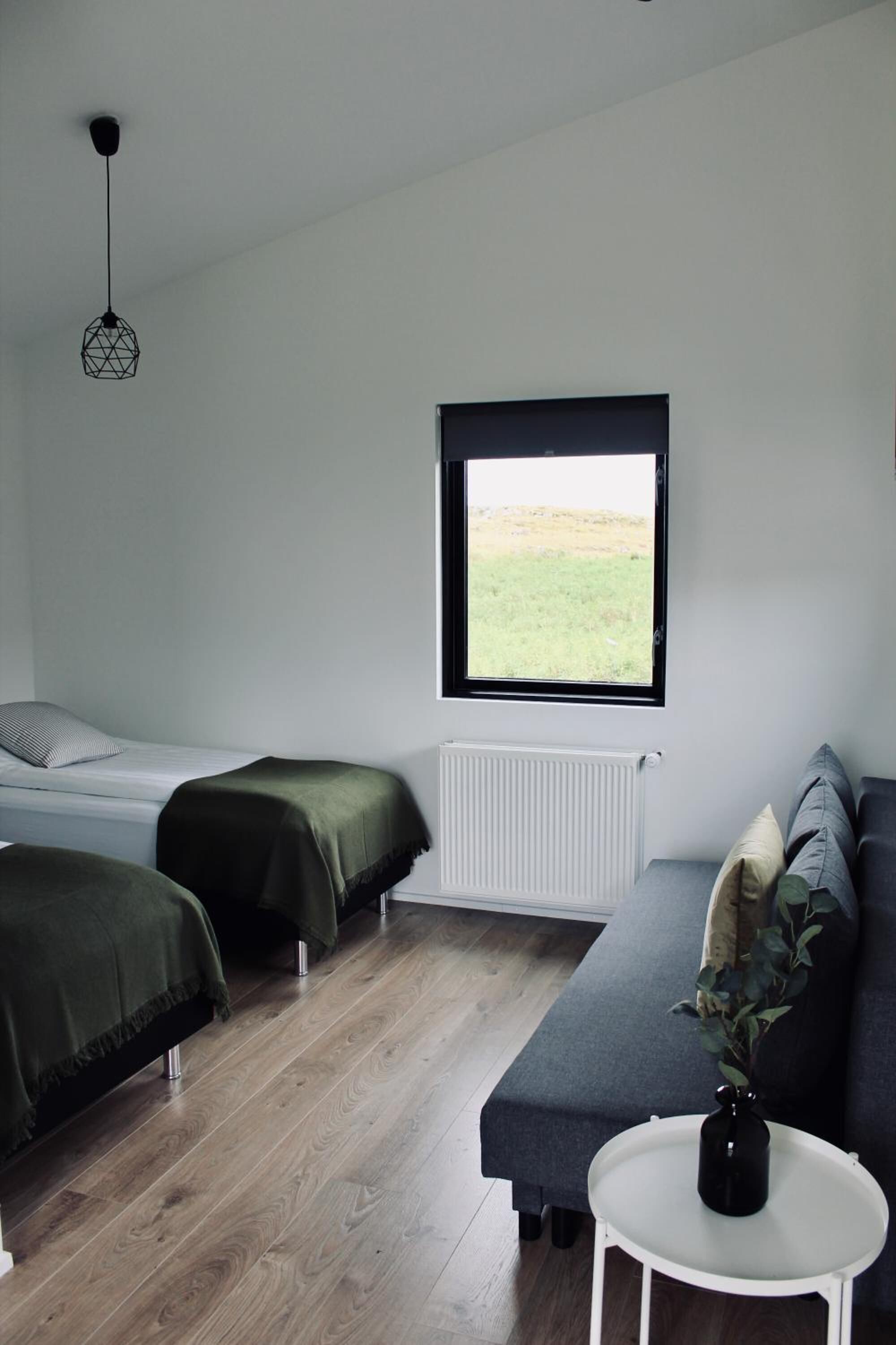 A bedroom with two beds, a window with a view, a radiator, and a small table with a vase.