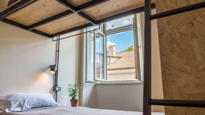 A single bed with a headboard, a bedside lamp, a potted plant, and a window with a view of buildings outside. - ControVento (Trieste)