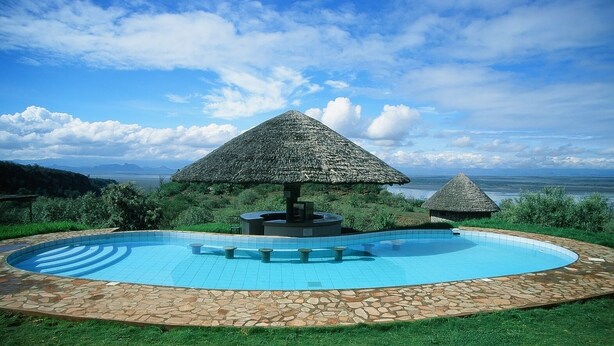 A swimming pool with a stone border, surrounded by grass and two thatched-roof structures. - Sunbird Lodge Elementaita (Nakuru)