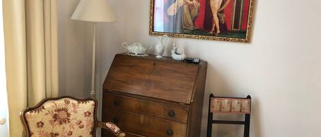A room with a floral-patterned armchair, a wooden dresser with drawers, a small chair, and a painting on the wall.