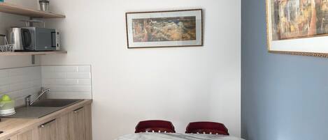 A compact kitchen with a sink, microwave, and a hanging blue chandelier. A framed painting hangs on the wall above a round table with four chairs.