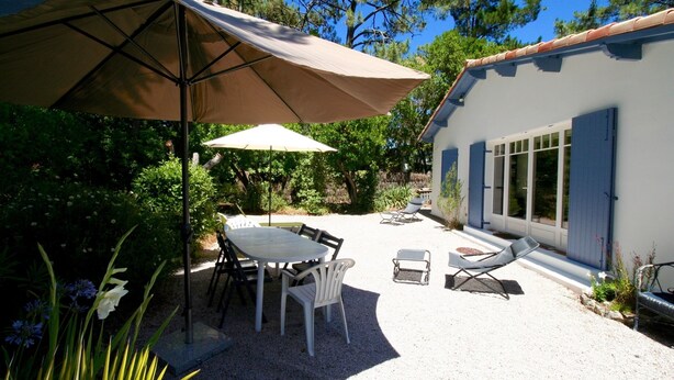 An outdoor patio with a table and chairs, a large umbrella, and a building with blue shutters. - Nice house with great garden in Cap Ferret, center + bay + ocean on foot (Lège-Cap-Ferret)