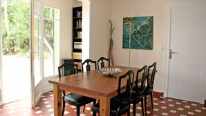 A dining area with a wooden table and chairs, a bookshelf, a painting, and a door. - Nice house with great garden in Cap Ferret, center + bay + ocean on foot (Lège-Cap-Ferret)