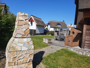 A stone wall, a thatched-roof building, a wooden deck with outdoor furniture, and a paved path. - Vacation home close to the beach with sauna, fireplace, washing machine & wifi incl. Incidental costs (Glowe)