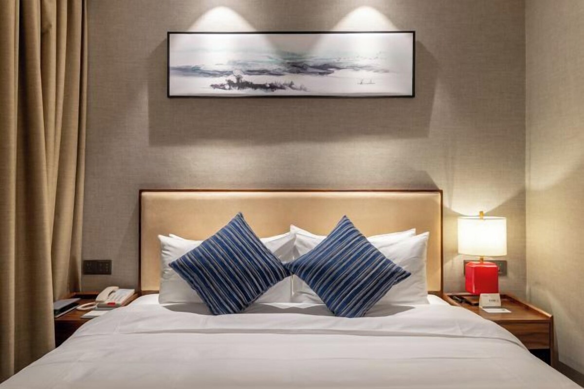 Habitación de hotel con cama, dos almohadas, mesita de noche con lámpara, teléfono y un cuadro en la pared.