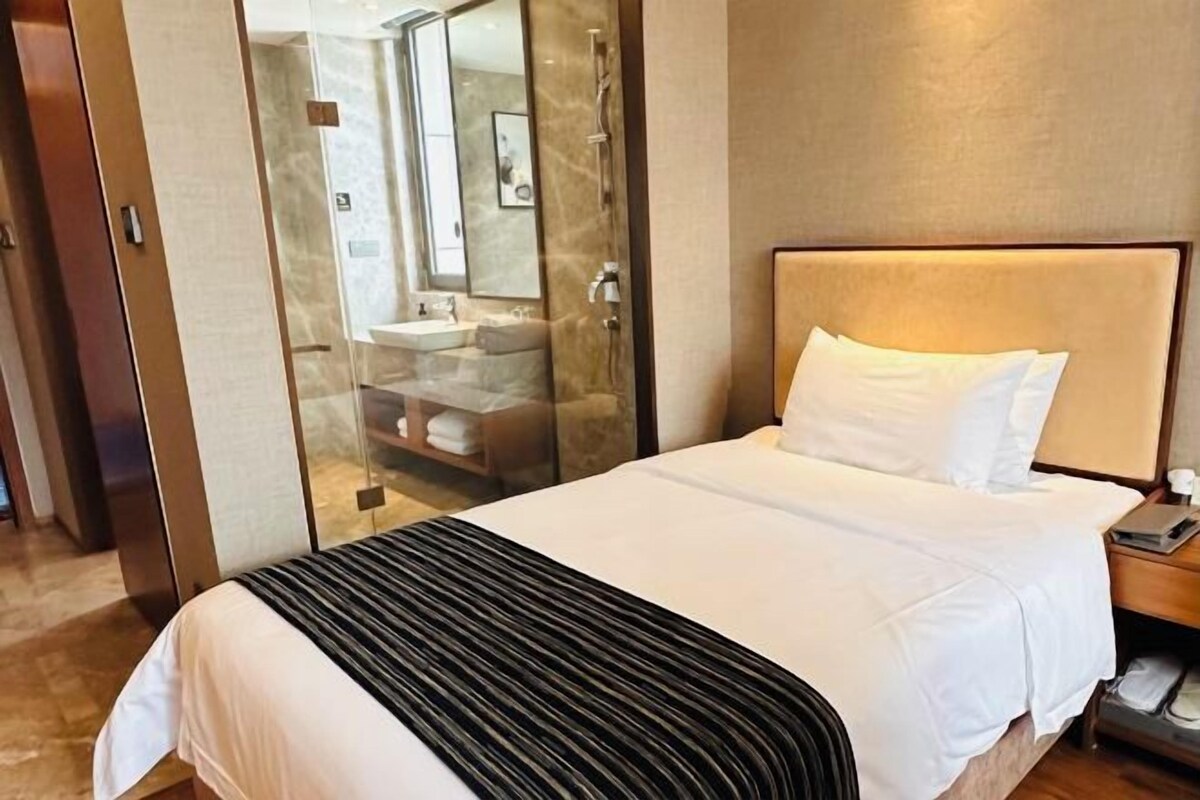 Una habitación de hotel con cama, mesita de noche con lámpara, cabina de ducha de vidrio y baño con lavamanos y espejo.